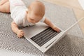 Indoor shot of charming little baby playing with laptop computer while lying on tummy on gray carpet, studying modern technology, Royalty Free Stock Photo