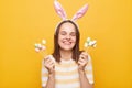 Indoor shot of charming positive attractive woman wearing bunny ears holding cake pops easter eggs isolated over yellow background Royalty Free Stock Photo