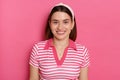 Indoor shot of cheerful charming young girl with hair band wearing striped casual t shirt, looking at camera with toothy smile, Royalty Free Stock Photo