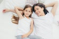 Indoor shot of cheerful mother and daughter embrace as lie on white bedclothes, wake up in morning, have weekend, being in