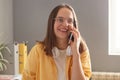 Indoor shot of cheerful woman wearing yellow shirt talking on cell phone, having pleasant conversation, sitting in office, Royalty Free Stock Photo