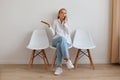 Indoor shot of confused helpless attractive female with ponytail sitting on chair against light wall, woman wearing stylish jeans