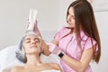 Indoor shot of cosmetologist applying to her client anti ageing mask with brush, holds big bottle of cream in hands. Lady keeps Royalty Free Stock Photo