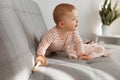 Indoor shot of curious charming infant baby girl wearing striped sleeper posing on gray sofa at home alone, looking away, finding Royalty Free Stock Photo