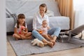 Indoor shot of cute baby, elder sister and young mother at home sitting on floor, woman wearing white shirt and jeans playing with Royalty Free Stock Photo