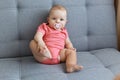 Indoor shot of cute beautiful baby daughter wearing pink bodysuit sitting on gray sofa with nipple in mouth, playing with leg, Royalty Free Stock Photo