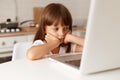 Indoor shot of cute bored dark haired female child sitting at table, looking at notebook screen with boring facial expression, Royalty Free Stock Photo