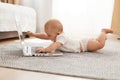 Indoor shot of cute curious baby girl or boy wearing white t shirt lying on floor on tummy, on gray carpet and touching laptop Royalty Free Stock Photo