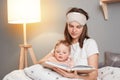 Indoor shot of dark haired woman posing with her daughter in bedroom, mother reading a book to baby while sitting together in bed Royalty Free Stock Photo