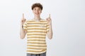 Indoor shot of delighted bright and happy pale guy with curly hairstyle and tattoos in striped yellow t-shirt raising