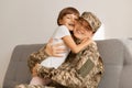 Indoor shot of delighted extremely happy military woman wearing camouflage uniform and cap posing with her daughter at home while Royalty Free Stock Photo