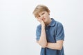 Indoor shot of displeased unhappy young boy with blond hair in blue t-shirt, making face palm and grimacing at camera Royalty Free Stock Photo