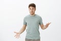 Indoor shot of displeased upset attractive guy in casual t-shirt, gesturing with spread palms and staring at camera