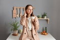 Indoor shot of dreamy young adult woman wearing beige suit standing near table on kitchen at home, holding cup in hand, enjoying