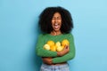 Indoor shot of emotional curly girl holding fresh oranges and lemons fruits, keeps mouth widely opened, dressed in