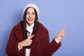 Indoor shot of energetic pleasant young woman wearing white sweater, hat and red fauxfur coat, making gesture, spending time alone