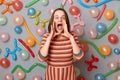 Indoor shot of extremely excited beautiful woman with brown hair wearing striped dress standing against gray wall with colorful