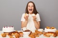 Indoor shot of extremely happy amazed woman with brown hair wearing white T-shirt sitting at table with cakes in hands, rejoicing Royalty Free Stock Photo