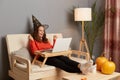 Indoor shot of extremely happy festive woman wearing witch hat working on laptop in home interior, clenched fists, celebrating