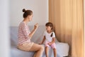 Indoor shot of female speech therapist working with little girl, people wearing casual style clothing, sitting on sofa near window