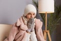 Indoor shot of freeze woman sitting on the sofa at home wearing winter coat, hat and mittens, warms hands in a cold house, looking Royalty Free Stock Photo