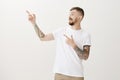 Indoor shot of friendly carefree caucasian fair-haired guy with tattoos and eartunnels, turining left and pointing up