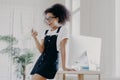 Indoor shot of happily surprised young unexperienced Afro American woman wears spectacles, white t shirt and sarafan, uses mobile