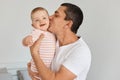 Indoor shot of happy brunette man wearing white casual t shirt holding his infant daughter and kissing her cheek, expressing great Royalty Free Stock Photo