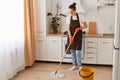 Indoor shot of happy Caucasian woman housewife cleaning floor with mop in kitchen, wearing white t shirt, jeans and brown apron, Royalty Free Stock Photo