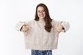 Indoor shot of happy charismatic young brunette female in glasses and sweater showing direction, pointing down withr