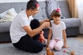 Indoor shot of happy family sitting on floor near gray couch, father makes hairstyle for his charming daughter, making pigtails, Royalty Free Stock Photo