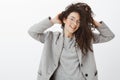 Indoor shot of happy fashionable female fashion blogger in stylih grey coat and glasses, touching curly hair and smiling Royalty Free Stock Photo