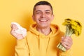 Indoor shot of happy man wearing casual style hoodie, holding dandelions and handkerchief in hands, suffering allergy but looking