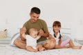 Indoor shot of happy positive family posing in light bedroom, young adult father sitting on bed with little daughters, infant and Royalty Free Stock Photo