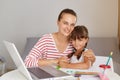 Indoor shot of happy positive woman with daughter, sitting at table with portable computer and books, female hugging her child, Royalty Free Stock Photo