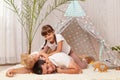 Indoor shot of happy smiling man wearing white t-shirt posing with his children on floor near wigwam, kids playing with daddy and