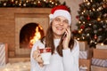 Indoor shot of happy smiling woman wearing white sweater and santa claus hat, sitting near fireplace and xmas tree, looking at Royalty Free Stock Photo