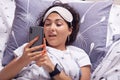 Indoor shot of happy young woman texting on mobile phone or having video call while lying in bed under blanket in morning, wearing Royalty Free Stock Photo