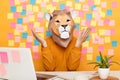 Indoor shot of helpless confused man in lion mask wearing orange sweater sitting at workplace with laptop, posing against yellow