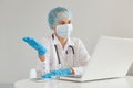 Indoor shot of helpless woman doctor wearing gown, surgical mask, medical cap and gloves, posing in front of notebook with spread