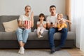 Indoor shot of impressed couple with children sitting on sofa at home in living room and watching shoking tv show or scary movie,