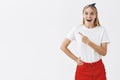 Indoor shot of impressed happy and intrigued attractive slim woman with blond hair in red skirt and headband pointing at