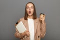 Indoor shot of inspired Caucasian brown-haired woman wearing jacket holding clipboard with documents and take away coffee,