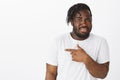 Indoor shot of intense unsure attractive plump guy with dark skin in white t-shirt, pointing left with index finger and