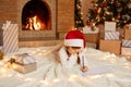 Indoor shot of little cute girl wearing white sweater and red hat, lying on floor on soft carpet in festive decorated room, Royalty Free Stock Photo