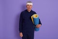 Indoor shot of mature man posing isolated over lilac background with yoga mat in hands, male wearing sports suit and head band, Royalty Free Stock Photo