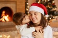 Indoor shot of mother and her little daughter hugging each other, having good mood, little cute girl kissing her mommy, merry