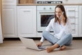 Indoor shot of optimistic dark haired woman wearing white t shirt and jeans sitting on floor in kitchen and talking phone while