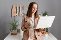 Indoor shot of optimistic joyful young adult woman freelancer wearing beige suit standing near table on kitchen, working on laptop