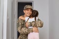 Indoor shot of optimistic man soldier wearing camouflage uniform and cap, posing with his daughter near the door, man going to Royalty Free Stock Photo